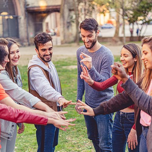 Eine Gruppe von Menschen verschiedener Ethnien stehen in einem Kreis draußen zusammen und halten ihre Hände in die Mitte. Sie scheinen etwas zu zählen.