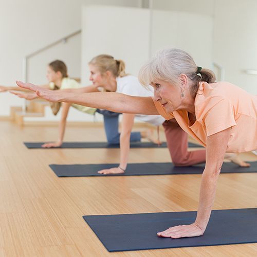 In einem hellen Gymnastikraum mit großem Spiegel knien drei Frauen unterschiedlichen Alters auf Matten. Dabei haben sie ein Bein nach hinten gestreckt und einen Arm nach vorne.