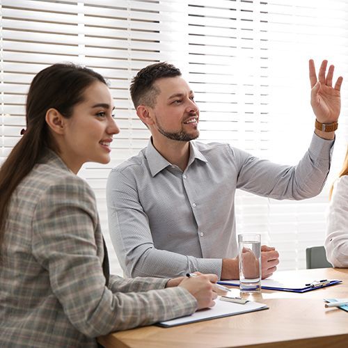 In einem hellen Raum stitzen an einem Holztisch mit Klemmbrettern zwei Personen. Zwischen zwei Frauen sitzt ein Mann der die Hand hebt. Im Hintergrund sind die weißen Jalousien eines großen Fensters zu sehen.