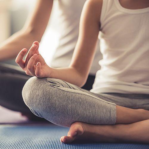 Auf Sportmatten sitzen zwei Frauen hintereinander im Lotussitz. Man sieht ihre Gesichter nicht. Sie tragen weiß-graue Sportkleidung.