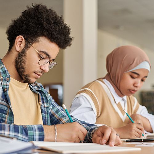 Zwei Personen of Colour sitzen an einem Tisch und schreiben etwas mit Bleistift auf Papier, das vor ihnen liegt.
