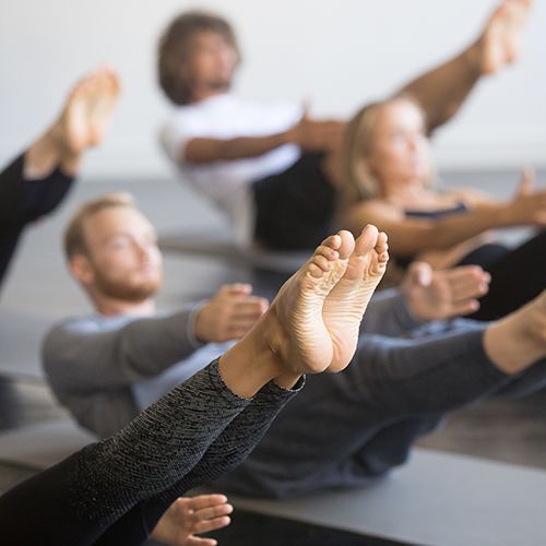 Ein Yoga-Raum mit dunklem Fußboden. Eine Gruppe von Menschen die auf dem Boden liegt und ihre Beine gestreckt nach vorne oben ausstreckt. Alle Personen sind undscharf. Ein paar Beine mit Füßen ist im Vordergrund im Fokus.