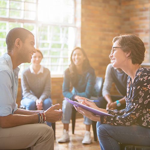 In einem sonnendurchflutetem Raum mit Backsteinmauer sitzen junge Menschen in einem Halbkreis zusammen. Eine Frau und ein Mann sitzen im Vordergrunud zu einem intensiverem Gespräch zusammen. Die Frau hält eine Mappe und einen Stift in den Händen.