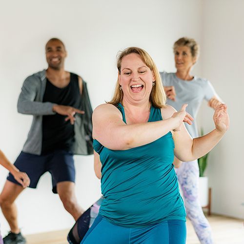 Mehrere Personen vielfältiger Herkunft in Sportkleidung haben Spaß bei einer dynamischen Übung. Im Fokus mittig ist eine Frau die ihre Arme rechts zur Seite schwingt und nach unten schaut. Der Raum ist hell und ein Bogenfenster ist zu sehen.
