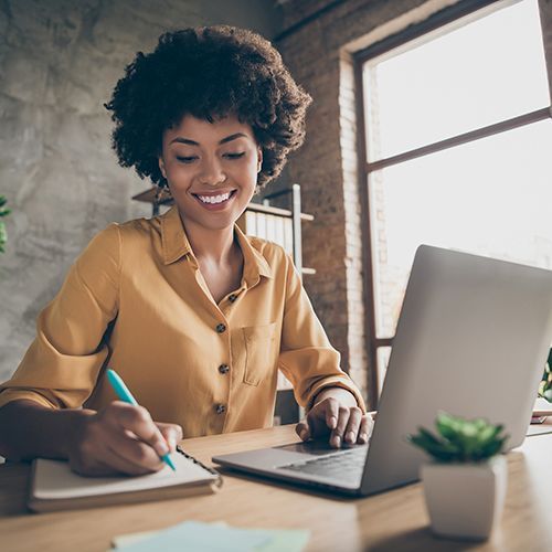 Eine Woman of Colour sitzt lächelnd an einem Tisch und hat vor sich einen Laptop und ein Notizbuch in das sie etwas notiert. Im Hintergrund ist eine rustikale Wand zwischen zwei Fenstern an denen bunte Post-Its kleben.