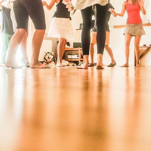 Junge Frauen und Männer stehen kreisförmig in einem großen Raum, halten sich an den Händen und tanzen miteinander.