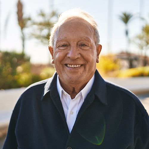 Ein älterer Mann schaut lächelnd in die Kamera. Er steht vor einem sonnigen hellen Hintergrund im freien. Es sind unscharf eine weiße Mauer und Pflanzen zu sehen.