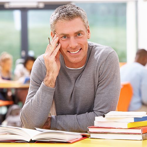 Das Foto zeigt einen Mann in einem modernen Klassenraum. Er sitzt an einem Tisch mit Büchern und lacht in die Kamera.