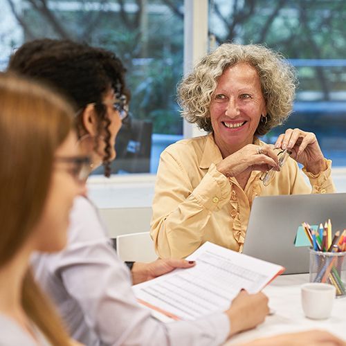 Drei Frauen sitzen an einem Tisch mit Laptop, Tablet und ein Glas mit Stiften. Zwei jüngere Frauen sind seitlich angeschnitten, eine ältere Frau schaut diese lachend an und hält ihre Brille in der Hand.