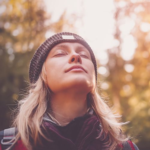 Eine Frau in Nahaufnahme. Sie trägt eine Mütze, hat die Augen geschlossen und reckt das Gesicht Richtung Himmel. Im Hintergrund sind Bäume im Abendlicht.
