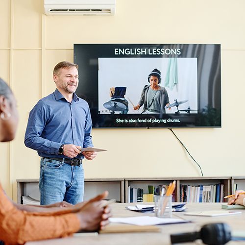 Ein Dozent steht in einem Seminarraum, hinter ihm ein Monitor mit einer Englisch-Lesson. Angeschnitten im Vordergrund einige Personne die zuhören.
